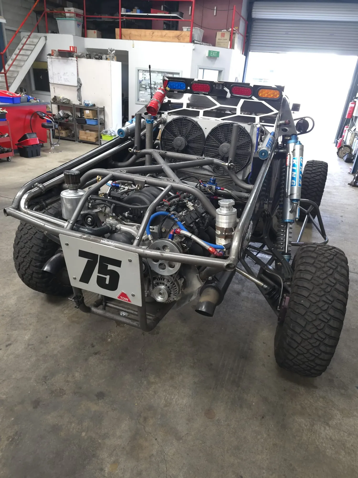 Number 75 tube-frame off-road race buggy in a workshop, showing the mid-mounted 6.0L LS V8, twin cooling fans and long-travel coilover suspension